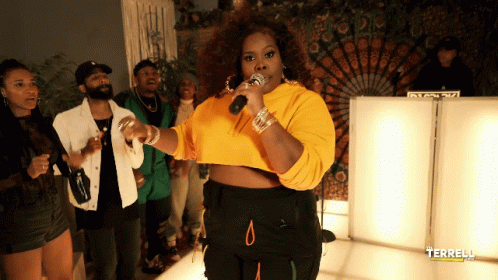 A Woman In A Yellow Crop Top Sings Into A Microphone In Front Of A Group Of People GIF