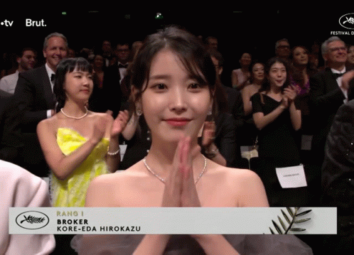 A Woman In A Yellow Dress Applauds In Front Of A Banner That Says Rang J Broker Kore-Eda Hirokazu GIF