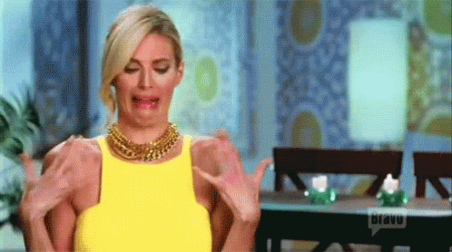 A Woman In A Yellow Dress Is Sitting At A Table With Her Arms Outstretched And Making A Funny Face . GIF