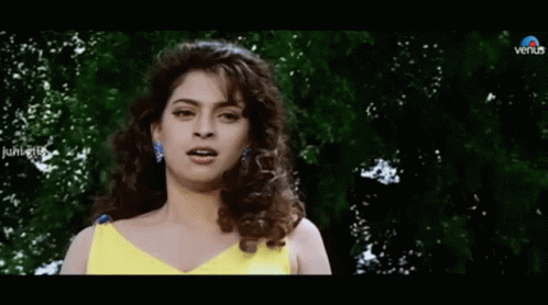 A Woman In A Yellow Dress Is Standing In Front Of Trees And Looking At The Camera . GIF