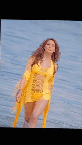 A Woman In A Yellow Dress Stands In Front Of A Body Of Water GIF