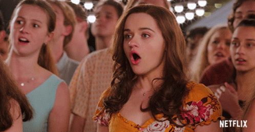A Woman In A Yellow Floral Dress Stands In A Crowd With Her Mouth Open And A Netflix Logo In The Background GIF