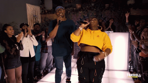 A Woman In A Yellow Hoodie Sings Into A Microphone In Front Of A Crowd GIF