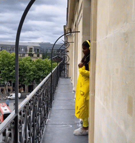 a woman in a yellow raincoat looks out from a balcony