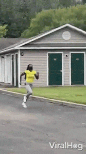 A Woman In A Yellow Shirt Is Running Down A Street In Front Of A House . GIF