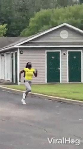 A Woman In A Yellow Shirt Is Running In Front Of A Building With Green Doors GIF