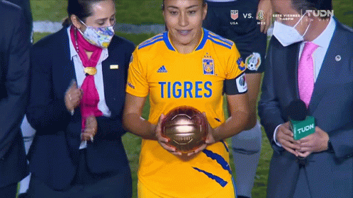 A Woman In A Yellow Shirt That Says Tigres Is Holding A Gold Ball GIF