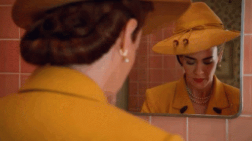 A Woman In A Yellow Suit And Hat Is Looking At Herself In A Mirror . GIF