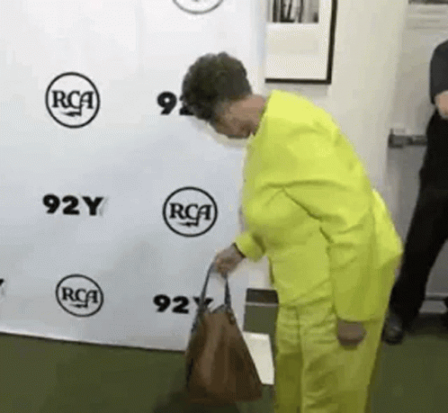 A Woman In A Yellow Suit Is Holding A Brown Purse In Front Of A Rca Sign . GIF