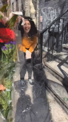 A Woman In A Yellow Sweater Is Standing In Front Of A Brick Wall With Graffiti On It GIF