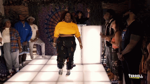 A Woman In A Yellow Sweatshirt Is Standing In Front Of A Group Of People With The Word Terrell On The Bottom Left GIF