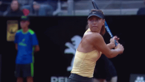 A Woman In A Yellow Tank Top Is Swinging A Tennis Racquet While A Man In A Blue Shirt Watches GIF