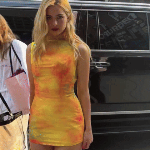 A Woman In A Yellow Tie Dye Dress Stands In Front Of A Black Van That Says Dsquared On The Side GIF
