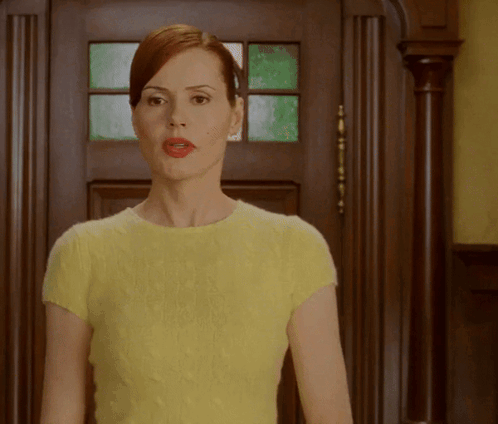 A Woman In A Yellow Top Is Standing In Front Of A Wooden Door GIF