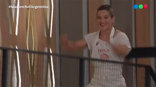 a woman in an apron is standing on a balcony with her arms outstretched and smiling .