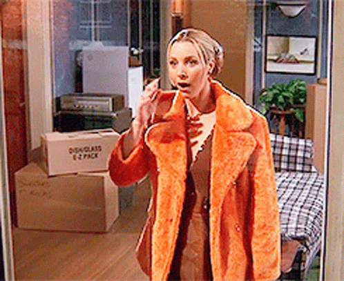 A Woman In An Orange Coat Is Talking On A Cell Phone In Front Of A Moving Box That Says 1 2 Pack GIF