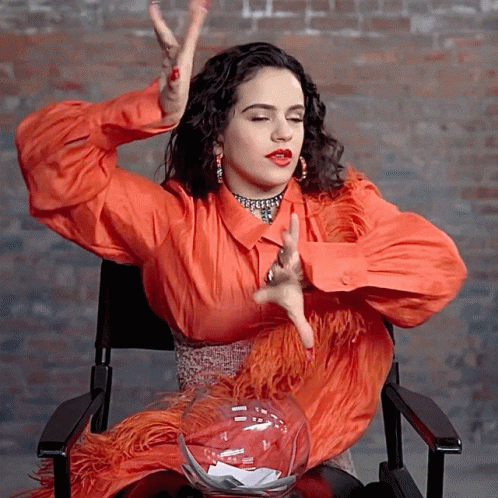 A Woman In An Orange Dress Is Sitting In A Chair GIF