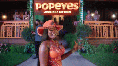 A Woman In An Orange Dress Is Standing In Front Of A Popeye 'S Louisiana Kitchen Sign GIF
