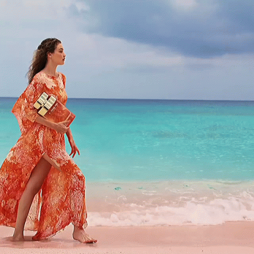 A Woman In An Orange Dress Walking On The Beach GIF