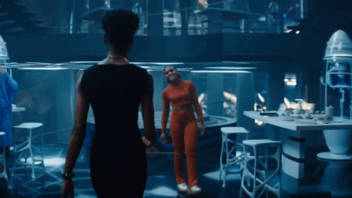 A Woman In An Orange Jumpsuit Stands Next To Another Woman In A Black Dress GIF
