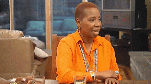 A Woman In An Orange Shirt Is Sitting At A Table With A Glass Of Water And A Watch . GIF