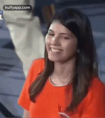 A Woman In An Orange Shirt Is Smiling And Looking At The Camera While A Man Stands Behind Her . GIF