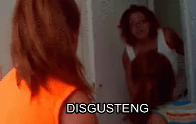 A Woman In An Orange Shirt Is Standing In Front Of A Mirror With The Word Disgusteng On It GIF