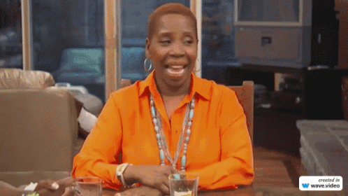 A Woman In An Orange Shirt Sits At A Table With Two Glasses Of Water GIF