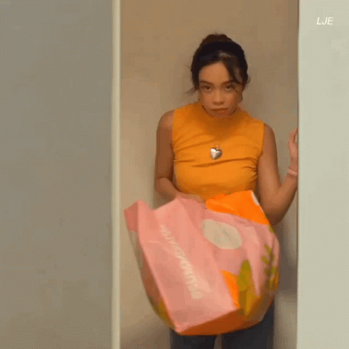 A Woman In An Orange Tank Top Is Standing In A Closet Holding A Pink And Orange Blanket . GIF