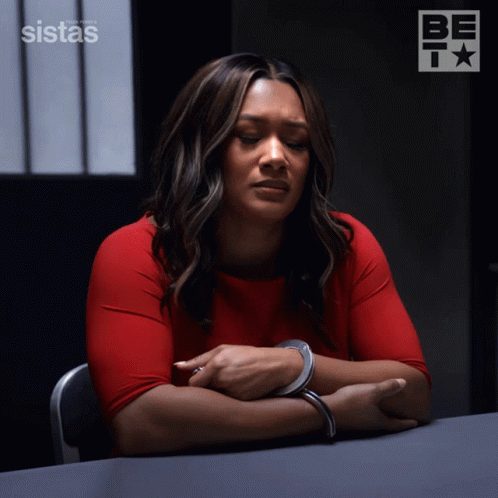 A Woman In Handcuffs Sits At A Table In Front Of A Sign That Says Sistas On It GIF
