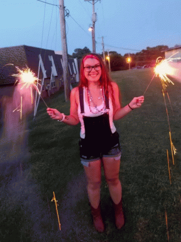 A Woman In Overalls Is Holding Sparklers In Front Of A Building That Says ' A ' On It GIF