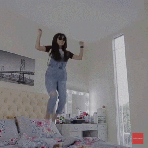 A Woman In Overalls Is Jumping In The Air In A Bedroom With A Picture Of A Bridge On The Wall GIF