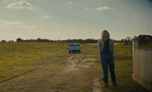 A Woman In Overalls Is Walking Down A Dirt Road With A Truck Behind Her GIF