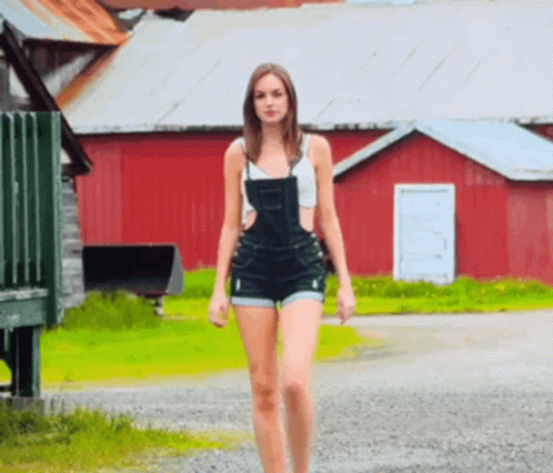 A Woman In Overalls Is Walking Down A Gravel Road In Front Of A Red Barn GIF