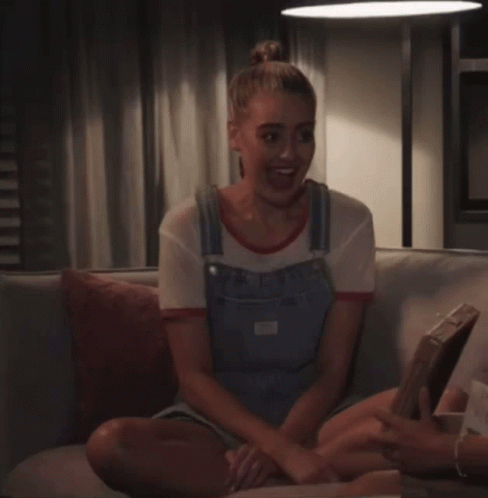 a woman in overalls sits on a couch while another woman holds a tray