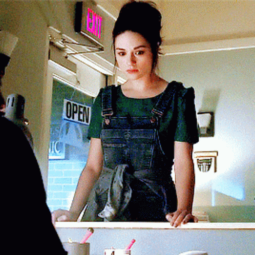 A Woman In Overalls Stands In Front Of A Sign That Says Open GIF