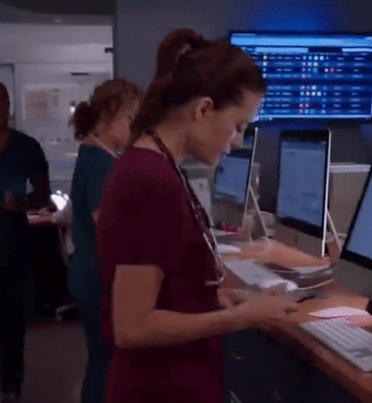 A Woman In Scrubs Is Looking At A Tablet In Front Of A Computer Monitor . GIF