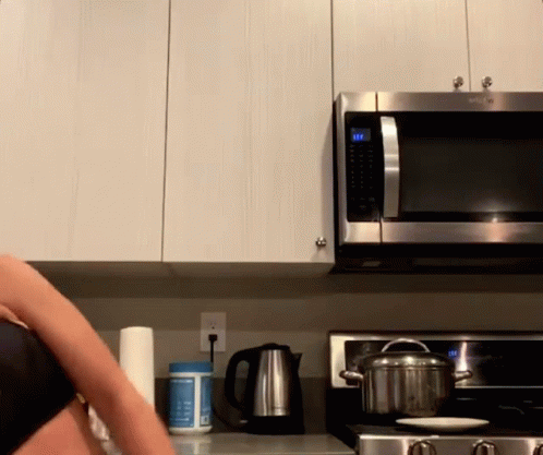 A Woman Is Cooking In A Kitchen With A Pot On The Stove And A Microwave That Has The Time Set At 10:22 GIF