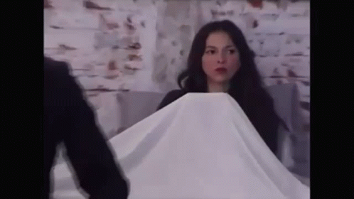 A Woman Is Covering Herself With A White Cloth While A Man Stands Behind Her . GIF