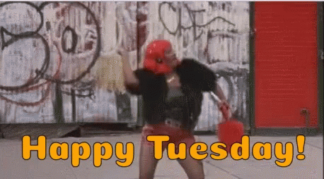 A Woman Is Dancing In Front Of A Graffiti Wall With The Words `` Happy Tuesday '' Written On It . GIF