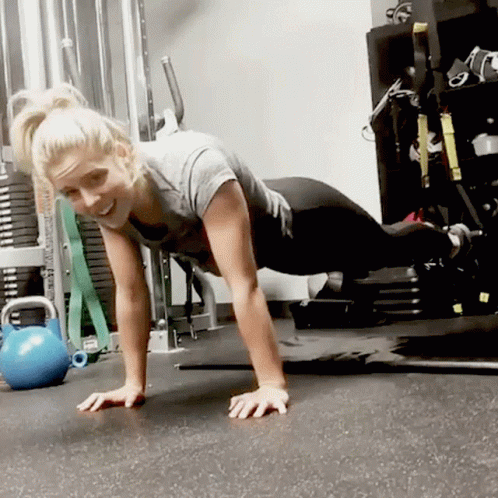A Woman Is Doing Push Ups In A Gym With A Blue Kettlebell In The Background GIF