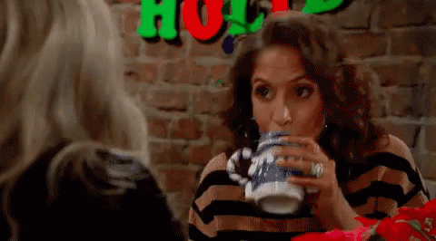 A Woman Is Drinking From A Mug While Sitting At A Table . GIF