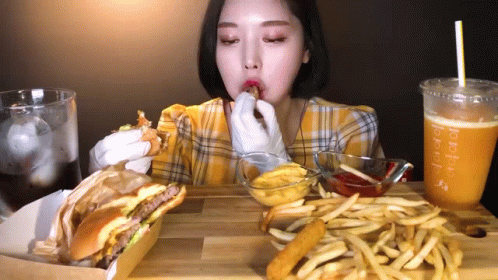 A Woman Is Eating A Hamburger And French Fries At A Table With A Drink That Says Tropical GIF