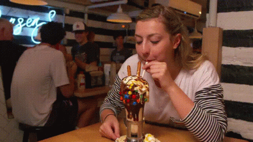 A Woman Is Eating A Milkshake With Pretzels And M & Ms On It GIF
