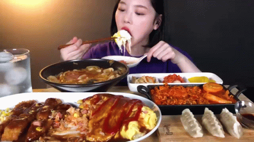 A Woman Is Eating A Plate Of Food With A Spoon And Chopsticks While Sitting At A Table GIF