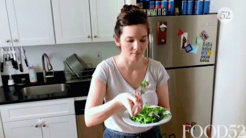 A Woman Is Eating A Salad In A Kitchen With A Sign That Says What It Is On The Refrigerator GIF