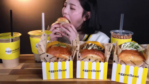 A Woman Is Eating A Sandwich In Front Of Boxes That Say Eggcellent On Them GIF