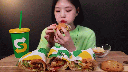 A Woman Is Eating A Subway Sandwich Next To A Cup That Says ' S ' On It GIF