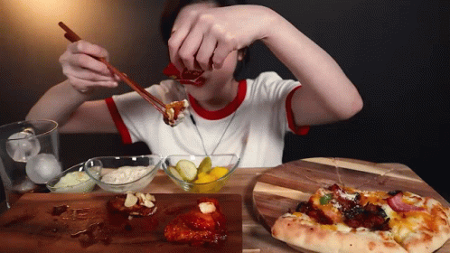 A Woman Is Eating Food With Chopsticks And A Pizza On A Wooden Cutting Board GIF