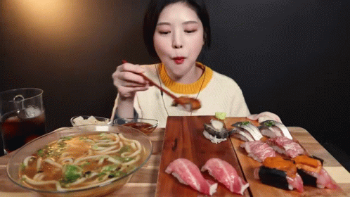 A Woman Is Eating Noodles And Sushi With Chopsticks At A Table GIF
